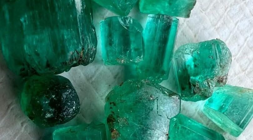 A close-up image of a vibrant, rough Nigerian emerald crystal, showcasing its natural form and rich green color. The emerald is unpolished and appears as it would be found in the mine, highlighting its authenticity.